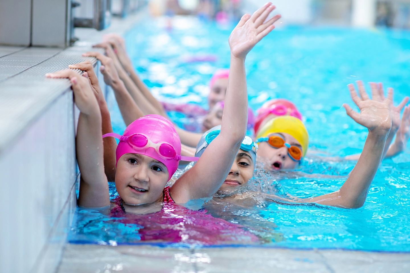 A group of children in brightly colored swim caps and goggles smile and wave while holding onto the edge of an indoor swimming pool. The clear blue water and playful atmosphere suggest a fun and engaging swim class for kids.