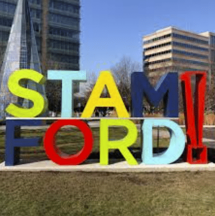 Large colorful letters spelling STAMFORD! stand outdoors in front of modern office buildings, with a lawn and trees visible in the background under a clear blue sky.